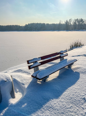 White shades of winter, a snow-covered bench by a frozen pond.の写真素材