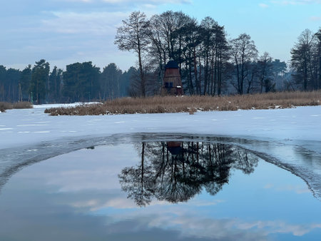 Reflection of trees and an old windmill in the unfrozen part of the water on the lake.の写真素材