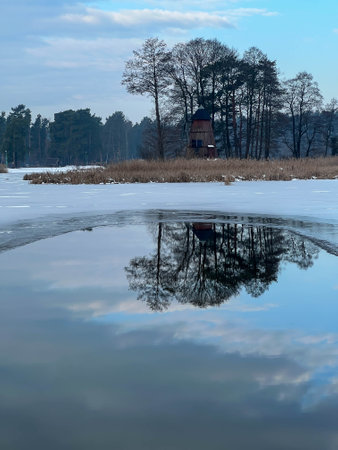 Reflection of trees and an old windmill in the unfrozen part of the water on the lake.の写真素材