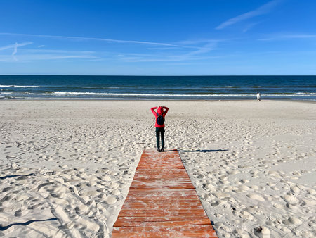 A young woman in a red jacket with a small backpack standing on planks looks out at the vastness of the sea, a beautiful beach of the Baltic Sea in Mrzezyno in May, Poland.の写真素材