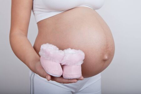 Young european pregnant woman hold in hand pink baby boots on grey background