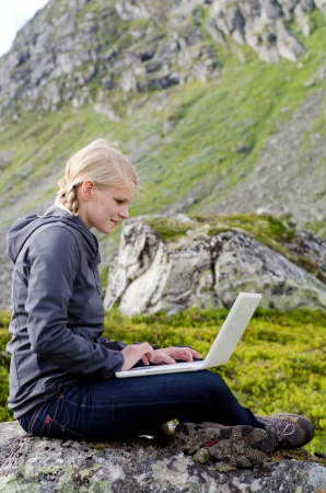 young blond woman sits with a laptop on a stone before mountain landscapeの写真素材