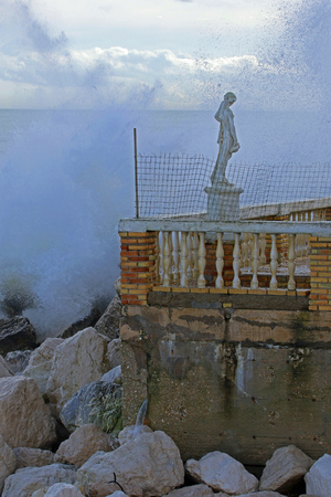 Rough sea in winter in Porto Recanati in the province of Ancona Italy near Monte Conero with the sea crashing against rocks and a statue on the terrace of a boarded up restaurantの写真素材