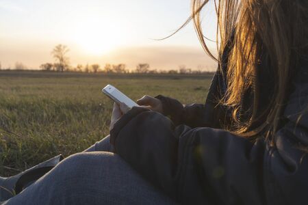 Girl uses a phone sitting in the field, sunsetの写真素材