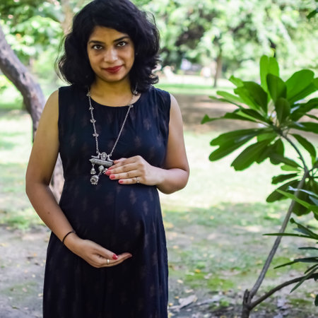 A pregnant Indian lady poses for outdoor pregnancy shoot and hands on belly, Indian pregnant woman puts her hand on her stomach with a maternity dress at society park, Pregnant outside maternity shootの写真素材