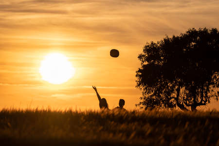 silhouette of girl and boy tossing hats in the air at a sunset in the countryside with the sun setting in the backgroundの写真素材