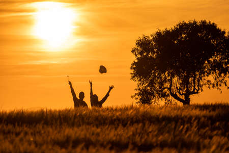 silhouette of girl and boy tossing hats in the air at a sunset in the countryside with the sun setting in the backgroundの写真素材