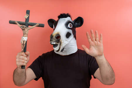 costumed person in a cow mask holding a crucifix simulating an exorcism with a pink background