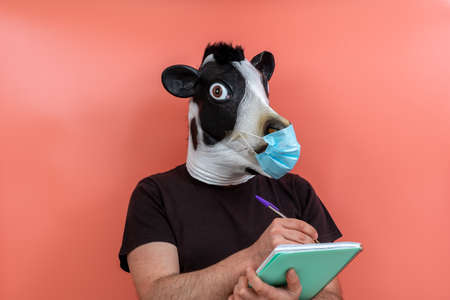 person wearing a cow mask and surgical mask taking notes with a pen on a notepad with pink backgroundの写真素材