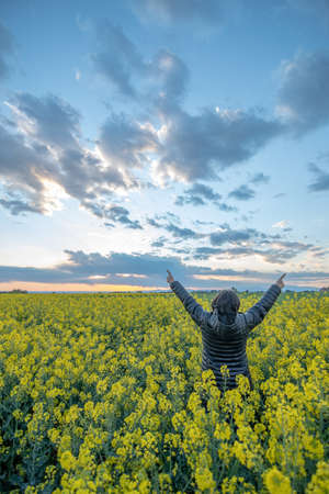 woman with her arms raised wearing a black anorak with the hood up in a field of yellow rapeseed...
