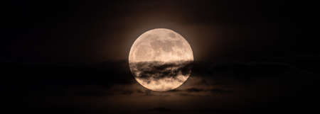 horizontal panoramic image of the bright full moon slightly obscured by clouds during the night.