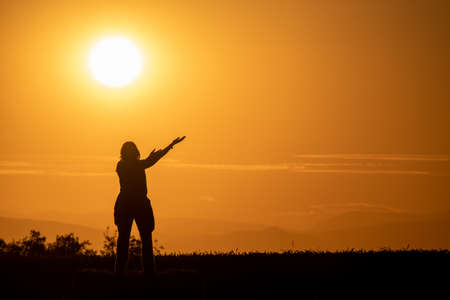 silhouette of a woman on a bale of straw in a field with her arms outstretched as if she were offering something with a large sun and mountains in the backgroundの写真素材