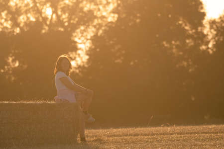 teenage girl with long blonde hair sitting on a straw bale in warm sunlight .Horizontal image of a rural sceneの写真素材