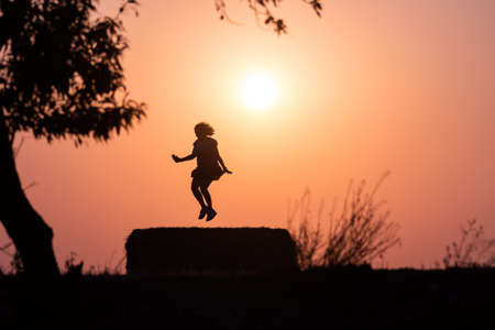 Silhouette of a latina woman jumping with the sun behind, in a warm sunset. Jumping over a bale of...