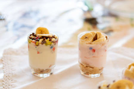Yogurt dessert with different fruits, crushed cookie and chocolate, presented in crystal glasses decorated with floral elements. Banana, almonds, cereals, chocolate chips. Natural and strawberry yogurt.の写真素材