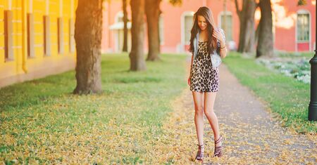 Stylish long hair brunette woman in a leopard animal print dress with sexy legs in brown high heel shoes, denim jacket, street fashion concept. Fashionable girl walking on summer street. Trendy style.の写真素材