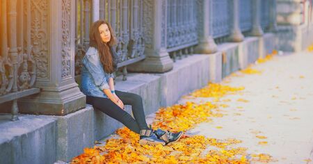 Young pretty hipster woman sitting near iron fence in a trendy denim jacket black jeans and fashion trendy boots. Beautiful millenial girl and yellow golden autumn maple leaves. Fall on city streetの写真素材