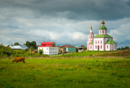 Suzdal - September 2017, Russia: Cow is grazing in a meadow, old residential houses, Church building and cloudy sky after the rainのeditorial素材
