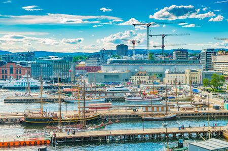 Oslo - June 2019, Norway: Aerial view of the harbor of Oslo with modern and historical ships. Cityscape with modern port, architecture, tower cranes, piers with walking people, hills and the blue skyのeditorial素材