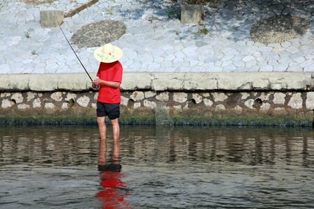 A Chinese fisherman with straw hat and red shirt on a riverbank