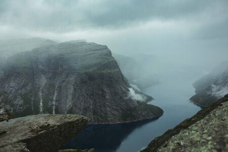 man on the edge of a troll tongue in norway in rainy weather on a background of mountains and a riverの写真素材