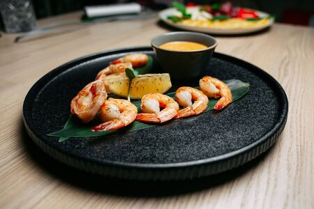 fried shrimp on a green leaf with a slice of orange and yellow sauce on a black plate on a wooden table backgroundの写真素材