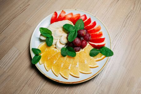 fruit sliced banana apple orange and grape with mint leaves on a white frosted plate on a wooden table backgroundの写真素材