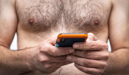 A man with a naked, hairy chest holds a mobile phone. Online chat, communication in apps, digital media website and social media.の写真素材