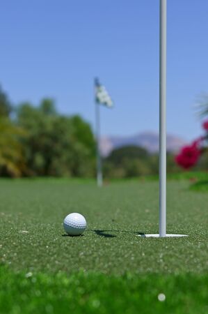 Golf ball on a putting green next to a hole.  The next hole is visible in the distance