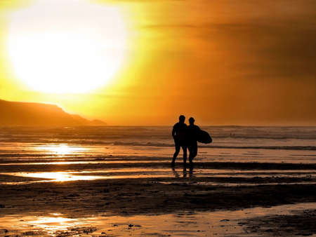Silhouette of a male and female surfer walking into the sea at sunset.の写真素材
