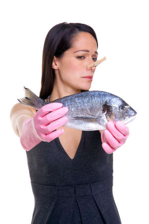 A woman holding a fish at arms length whilst wearing pink rubber gloves and a clothes peg on her nose. Isolated on white background, focus on the fish.の写真素材