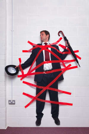 Businessman holding an umbrella and bowler hat stuck to a wall with red tape.