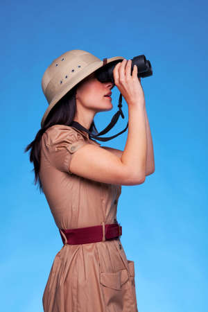 A woman wearing a pith helmet searching with a pair of binoculars, blue background with copy space.の写真素材