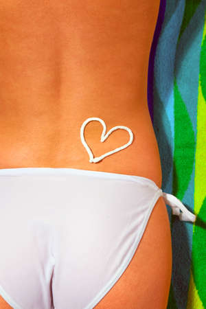 Lower half of a woman sunbathing wearing white bikini bottoms with a suntan cream drawing of a heart on her backの写真素材