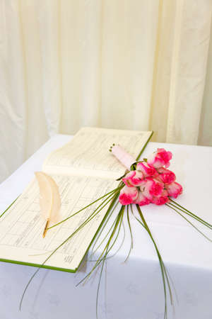 A wedding register and quill with a bouquet of pink roses on a white table.の写真素材