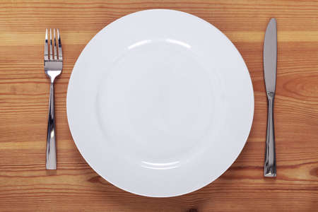 Photo of an empty white plate with knife and fork on a rustic wooden table.の写真素材
