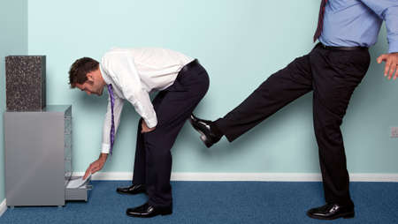 Photo of a businessman bending over to get something out of a drawer as a colleague kicks him up the backside.の写真素材