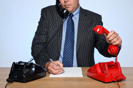 Photo of a businessman sat at a desk with two traditional telephones, one red and one black. He is listening to one call whilst picking up the other phone.の写真素材