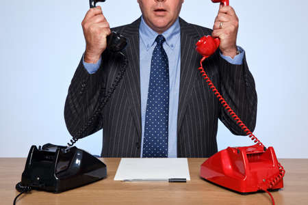 Photo of a businessman sat at a desk with two traditional telephones, one red and one black. Both handsets are raised and he doesn't know who to talk to first.の写真素材