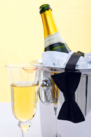 Photo of a bottle of chilled Champagne in an ice bucket with a black bow tie drapped over it and a glass with a red lipstick mark on.の写真素材