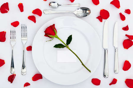 Photo of a table place setting with a red rose and blank card on the plate plus rose petals on the tablecloth. Add your own message to the place card.の写真素材