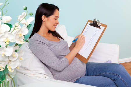 Photo of a pregnant woman at home sitting in an armchair writing a list of possible boy and girl names for her baby.の写真素材