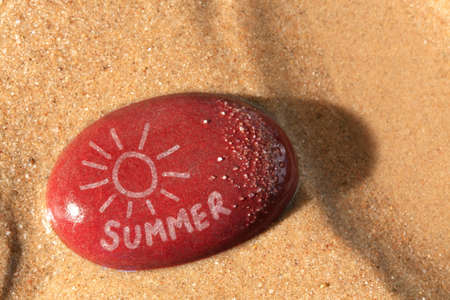 Concept photo of a wet red pebble on a sandy beach with a drawing of the sun and the word summer on it with the late setting sunshine casting a shadow across the sand.の写真素材