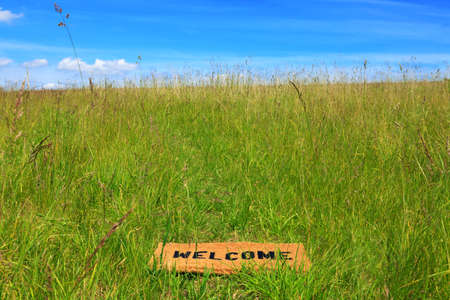 Photo of a welcome doormat in a grass meadow on a bright sunny day with blue sky and sunshine.の写真素材