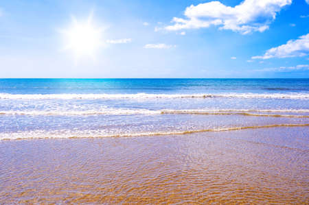 Photo of waves crashing on a golden beach with the sun shining in a blue sky の写真素材