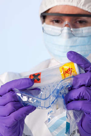 Photo of a forensic lab technician in protective workwear examining a plastic container that has white powder in and is marked biohazard and irritant inside a police evidence bag.の写真素材