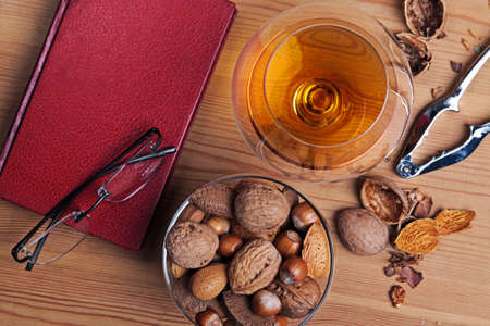 Still life photo of cognac in a crystal brandy glass with book, reading glasses and a selection of nuts on a table.の写真素材