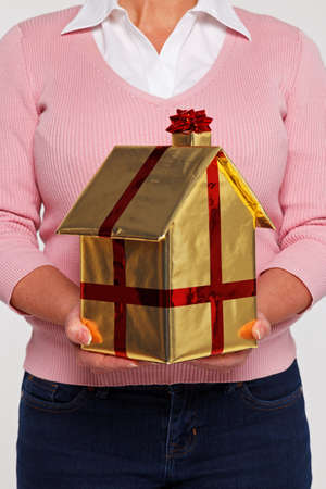 New home concept image of a woman in casual clothing holding a house gift wrapped in gold paper with red ribbon and bow.の写真素材