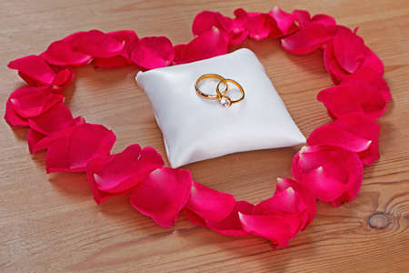 Gold wedding and engagement ring on a small white cushion surrounded by a rose petal heart on a wooden background.の写真素材