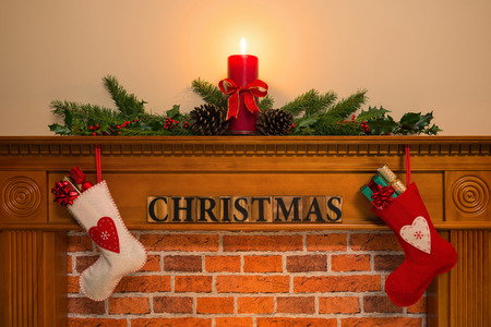 Mantelpiece with red candle and fresh garland made from holly, two stockings full of gifts hanging over the fireplace with the word Christmas.の写真素材
